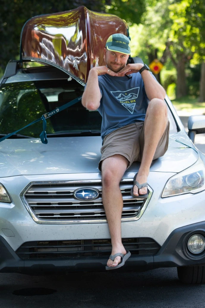 A man posing on the hood of a car wearing a Level Six T-shirt with a canoe in the background.